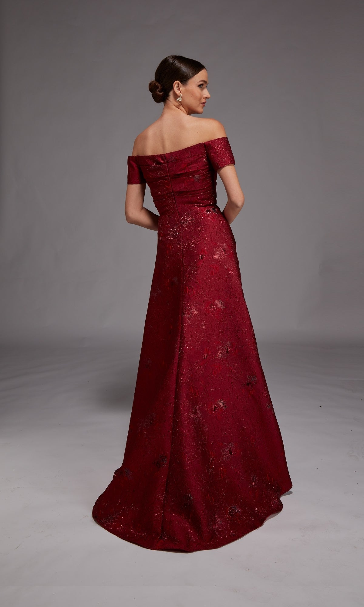 A woman stands facing away in the Alyce 27828 long formal dress, showcasing an off-the-shoulder design and subtle floral jacquard fabric, set against a studio backdrop.